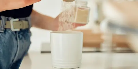 Osoba wsypuje łyżeczką słodzik do białego kubka.