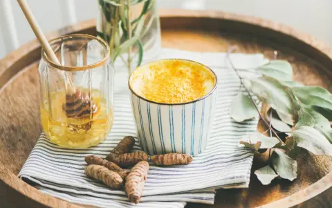 Aromaticzne złote mleko w kubeczku, obok słoiczek z miodem i korzenie kurkumy. Idealne na chłodne wieczory.