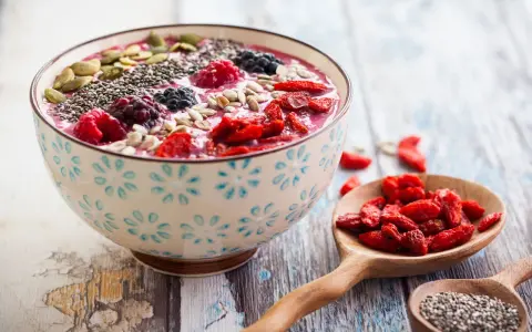 Miseczka smoothie z owocami, nasionami chia, pestkami dyni i słonecznika. Obok łyżka z jagodami goji. Nasiona chia jak jeść? Dodaj do smoothie!